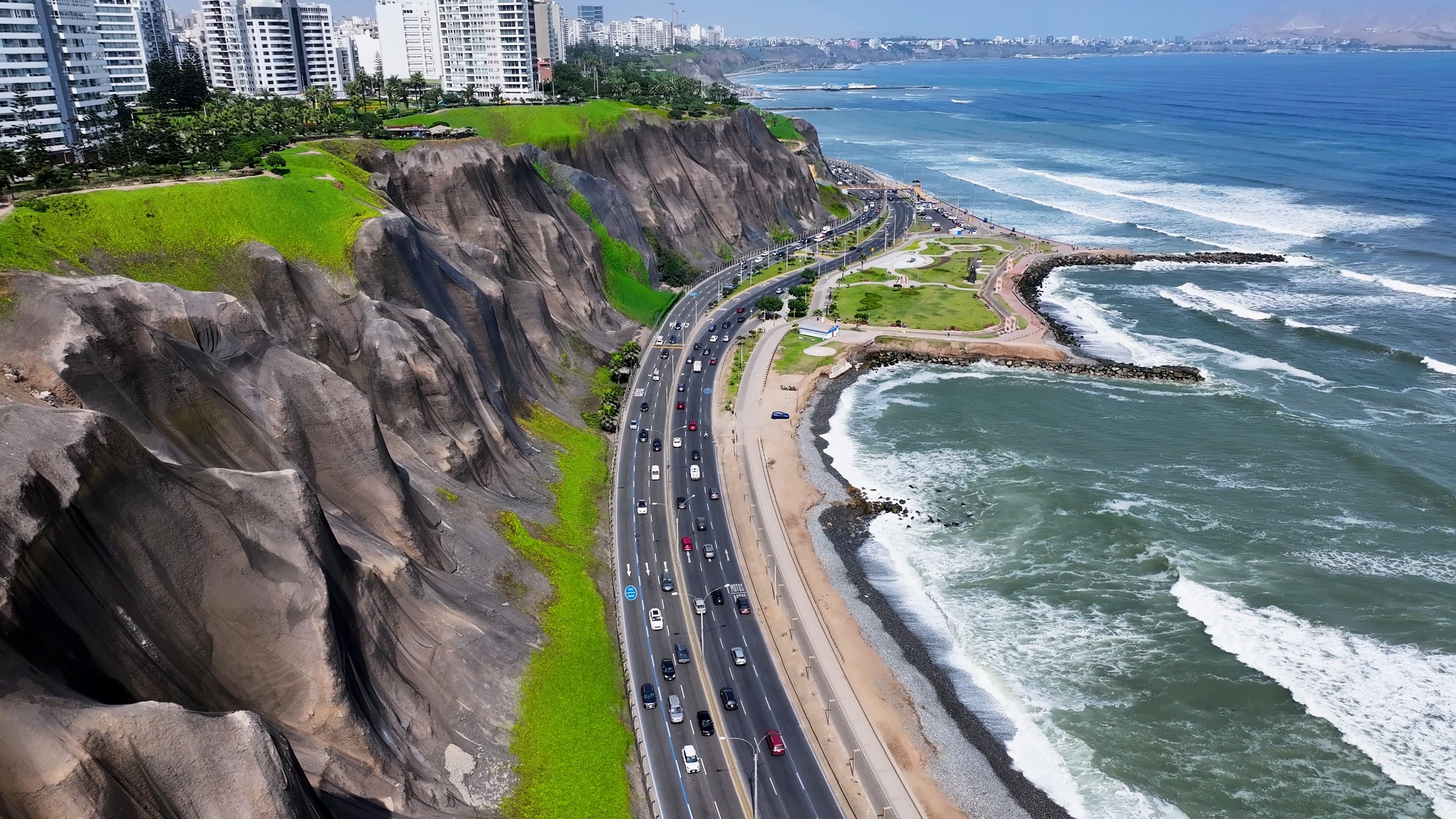 Vista aérea de acantilados, una autopista costera con vehículos y el océano con olas blancas.